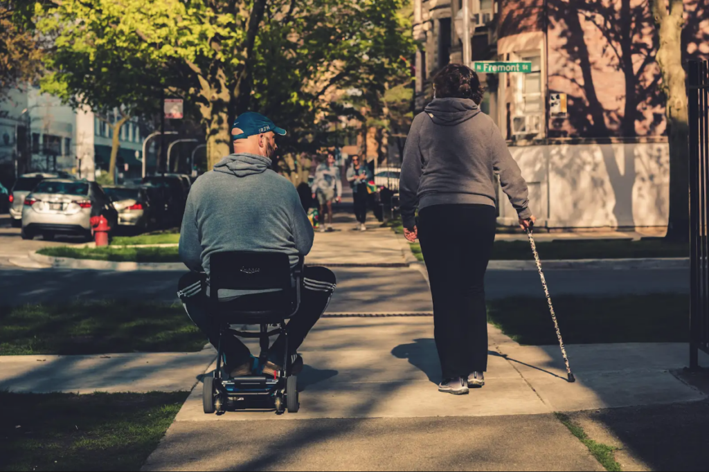 Two people going down a sidewalk, both using mobility devices
